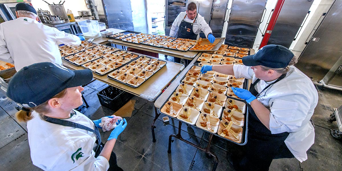 Four chefs, surrounding tables, fill trays while preparing a food course for a gala event