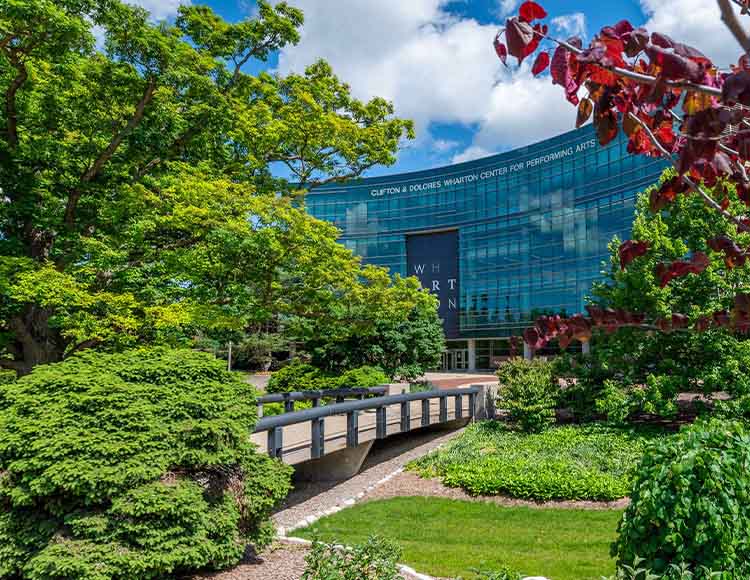 A bridge among greenery and trees in front of the Wharton Center of Performing Arts.