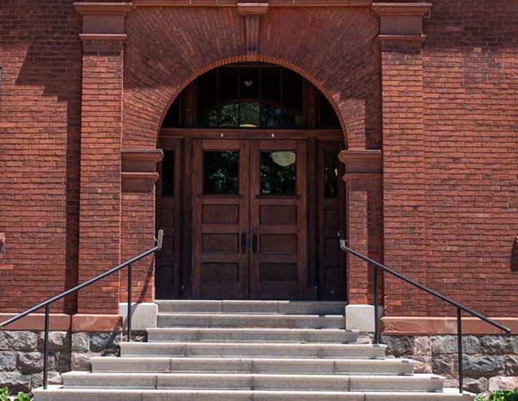 An exterior shot of a red brick building’s arched doorway. There are concrete steps and metal railings leading to the wooden doors.