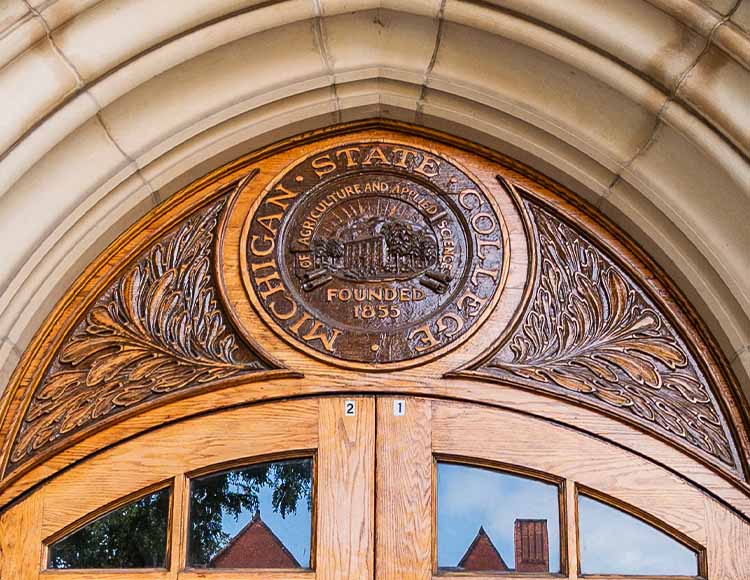 An ornate wooden carving at the top of a door that says Michigan State College of Agriculture and Applied Sciences founded 1855.