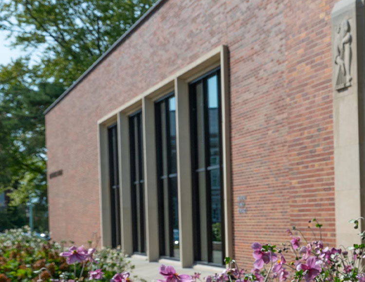 A brick building with four vertical windows and pink and purple flowers in the foreground.