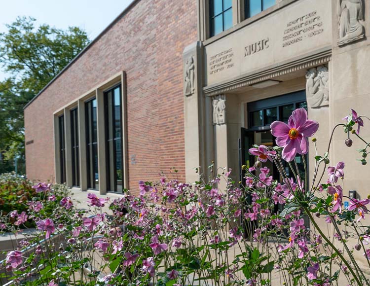 A brick building with four vertical windows and pink and purple flowers in the foreground.